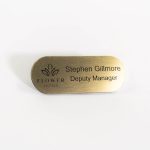 A pill shaped brushed gold with black engraved text, with logo of flowers hotels logo on the left.