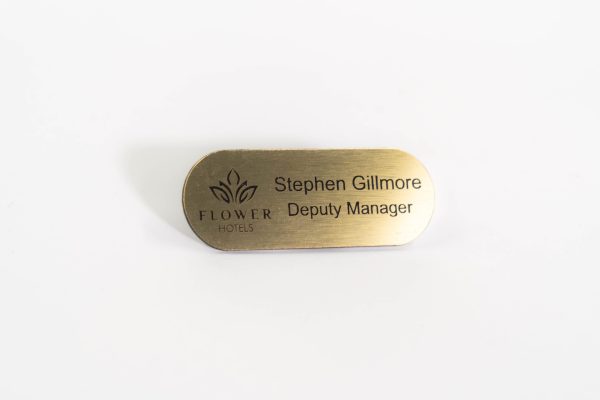 A pill shaped brushed gold with black engraved text, with logo of flowers hotels logo on the left.