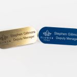 Pill shaped brushed gold name badge with black engraved text, Also a navy blue with engraved white text and flowers hotel logo on the left