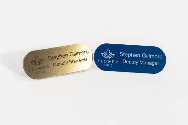 Pill shaped brushed gold name badge with black engraved text, Also a navy blue with engraved white text and flowers hotel logo on the left
