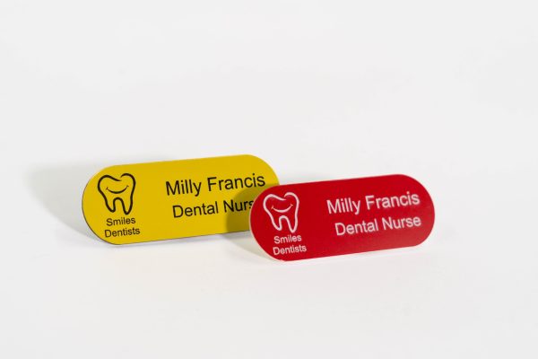 A red pill shaped name badge with white engraved text also a yellow pill shaped badge with black text and smiles dental logo on the left