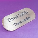 A polished brass badge with a pill-like shape, featuring black engraved text