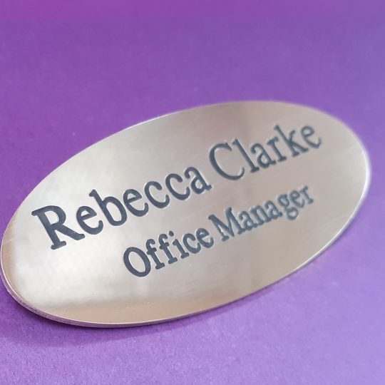 Polished brass badge in the shape of an oval, featuring black engraved text