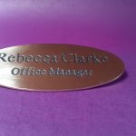 Polished brass badge in the shape of an oval, featuring black engraved text