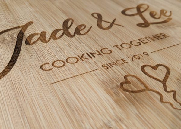 Close up of a bamboo chopping board showcasing 'Jade & Lee Cooking Together Since 2019' and two lovely love hearts.