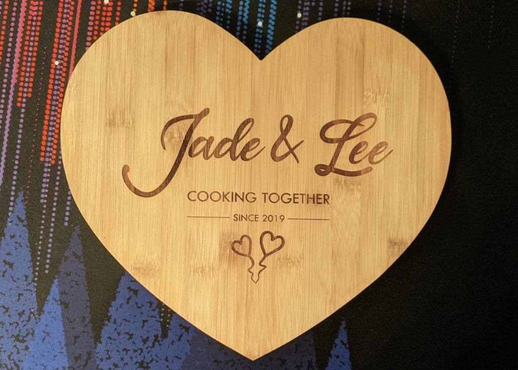 Bamboo heart-shaped chopping board with custom engraving: 'Jade & Lee Cooking Together Since 2019' and two love hearts.