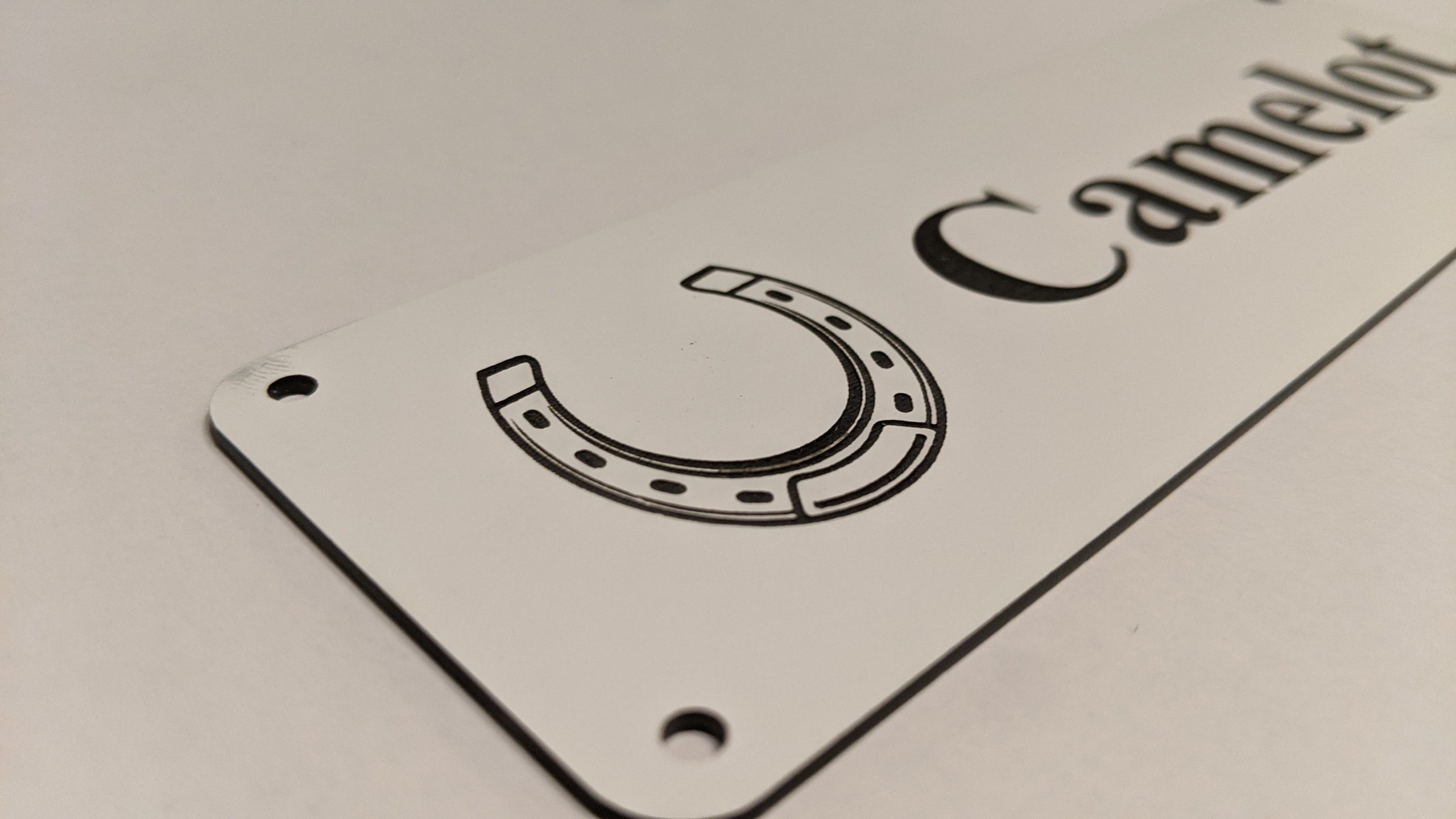 Close up of black engraving on white Stable Name Plaque – 6″ x 2″ with Left-Hand Horseshoe Design