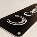 Close up of white engraving on black Stable Name Plaque - 6" x 2" with Left-Hand Horseshoe Design