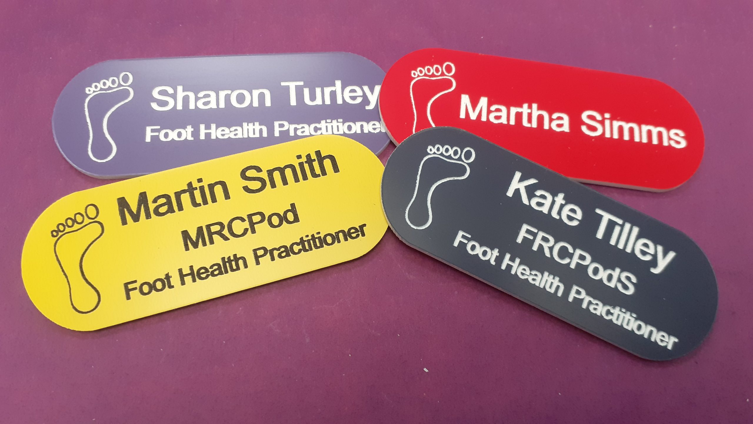 Red, yellow, purple, navy blue name badge in the shape of a pill with foot logo and employee name and title engraved in black or white text