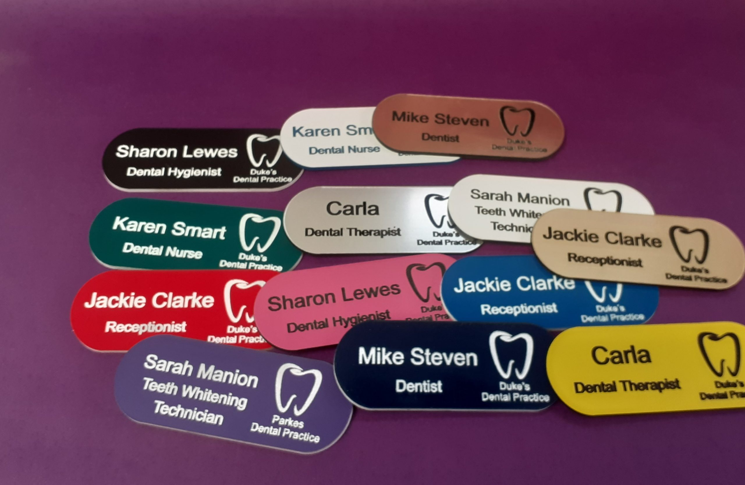 A collection of vibrant and diverse name badges, each uniquely coloured in shades such as blue, red, green, and yellow. The badges are designed in the shape of a pill, featuring a distinctive tooth logo on the left-hand side
