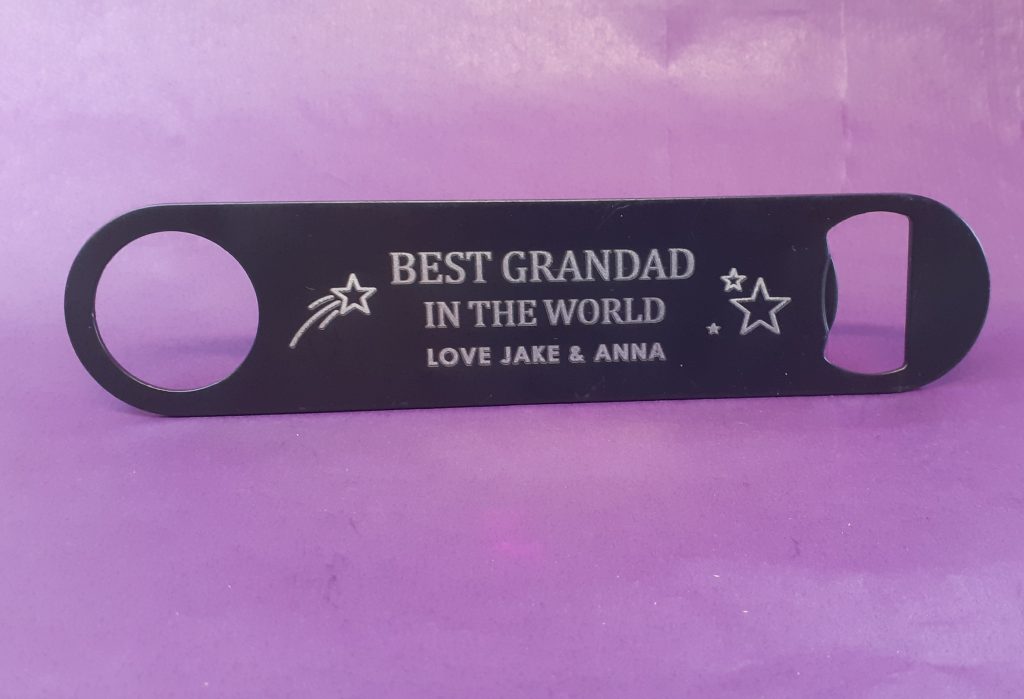 black metal bottle opener. It has a personalised message engraved on it that says: BEST GRANDAD IN THE WORLD LOVE JAKE & ANNA There are also some star designs on it. The bottle opener is against a purple background.