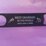 black metal bottle opener. It has a personalised message engraved on it that says: BEST GRANDAD IN THE WORLD LOVE JAKE & ANNA There are also some star designs on it. The bottle opener is against a purple background.