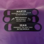 black metal bottle opener. It has a personalised message engraved on it that says: BEST GRANDAD IN THE WORLD LOVE JAKE & ANNA There are also some star designs on it. The bottle opener is against a purple background.