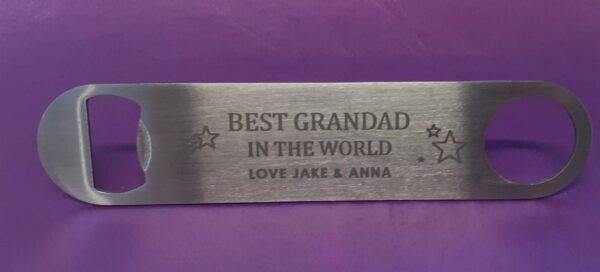 steel metal bottle opener. It has a personalised message engraved on it that says: BEST GRANDAD IN THE WORLD LOVE JAKE & ANNA There are also some star designs on it. The bottle opener is against a purple background.
