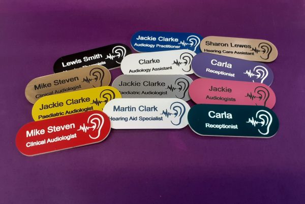 A collection of vibrant and diverse name badges, each uniquely coloured in shades such as blue, red, green, and yellow. The badges are designed in the shape of a pill, featuring a distinctive hearing logo on the right-hand side