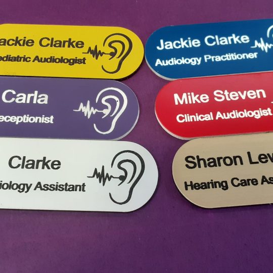 A collection of vibrant and diverse name badges, each uniquely coloured in shades such as blue, red, green, and yellow. The badges are designed in the shape of a pill, featuring a distinctive hearing logo on the right-hand side