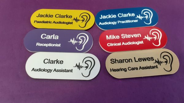 A collection of vibrant and diverse name badges, each uniquely coloured in shades such as blue, red, green, and yellow. The badges are designed in the shape of a pill, featuring a distinctive hearing logo on the right-hand side