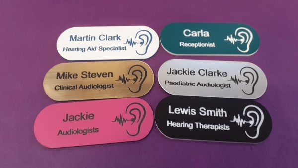 A collection of vibrant and diverse name badges, each uniquely coloured in shades such as blue, red, green, and yellow. The badges are designed in the shape of a pill, featuring a distinctive hearing logo on the right-hand side