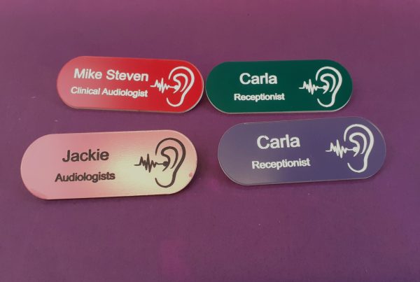 A collection of vibrant and diverse name badges, each uniquely coloured in shades such as blue, red, green, and yellow. The badges are designed in the shape of a pill, featuring a distinctive hearing logo on the right-hand side