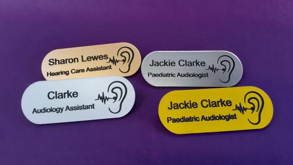 A collection of vibrant and diverse name badges, each uniquely coloured in shades such as blue, red, green, and yellow. The badges are designed in the shape of a pill, featuring a distinctive hearing logo on the right-hand side