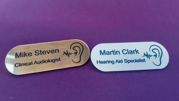 A collection of vibrant and diverse name badges, each uniquely coloured in shades such as blue, red, green, and yellow. The badges are designed in the shape of a pill, featuring a distinctive hearing logo on the right-hand side
