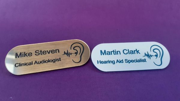 A collection of vibrant and diverse name badges, each uniquely coloured in shades such as blue, red, green, and yellow. The badges are designed in the shape of a pill, featuring a distinctive hearing logo on the right-hand side