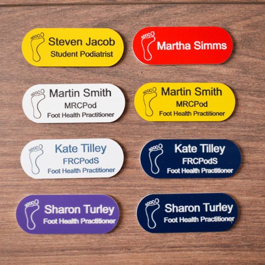 A collection of vibrant and diverse name badges, each uniquely coloured in shades such as blue, red, green, and yellow. The badges are designed in the shape of a pill, featuring a distinctive hearing logo on the right-hand side