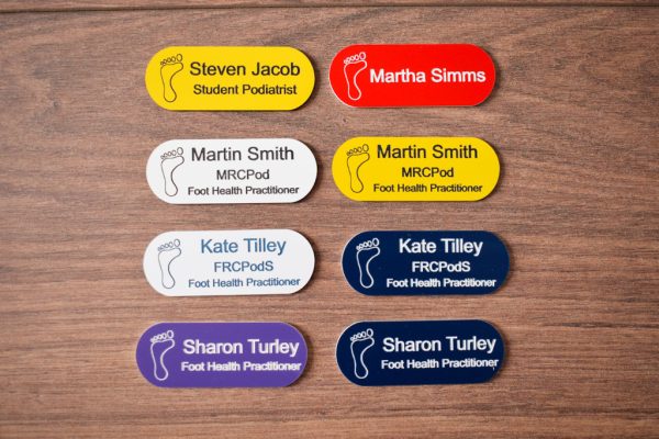 A collection of vibrant and diverse name badges, each uniquely coloured in shades such as blue, red, green, and yellow. The badges are designed in the shape of a pill, featuring a distinctive hearing logo on the right-hand side