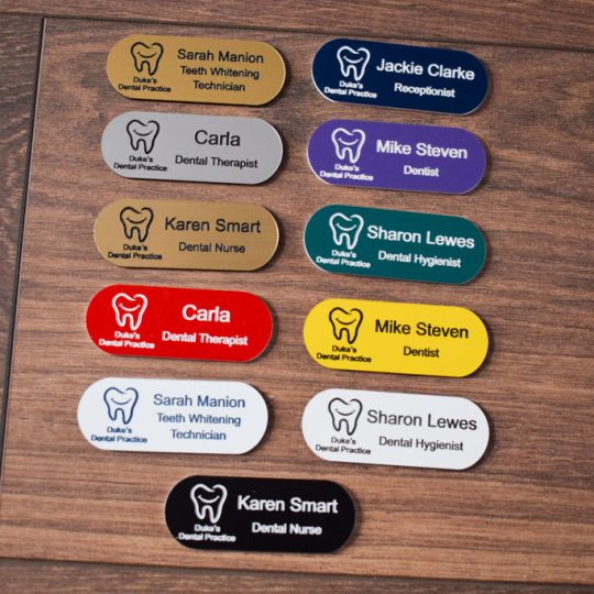 A collection of vibrant and diverse name badges, each uniquely coloured in shades such as blue, red, green, and yellow. The badges are designed in the shape of a pill, featuring a distinctive hearing logo on the right-hand side