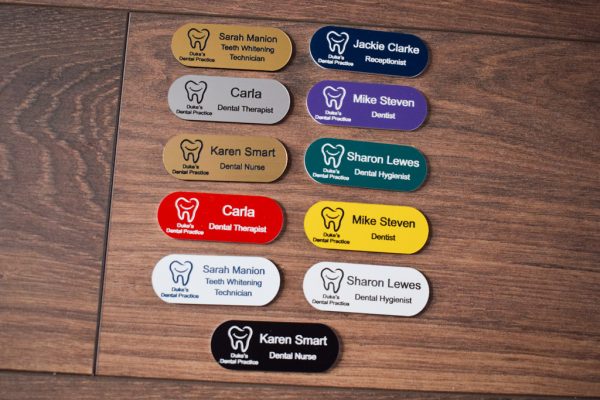 A collection of vibrant and diverse name badges, each uniquely coloured in shades such as blue, red, green, and yellow. The badges are designed in the shape of a pill, featuring a distinctive hearing logo on the right-hand side