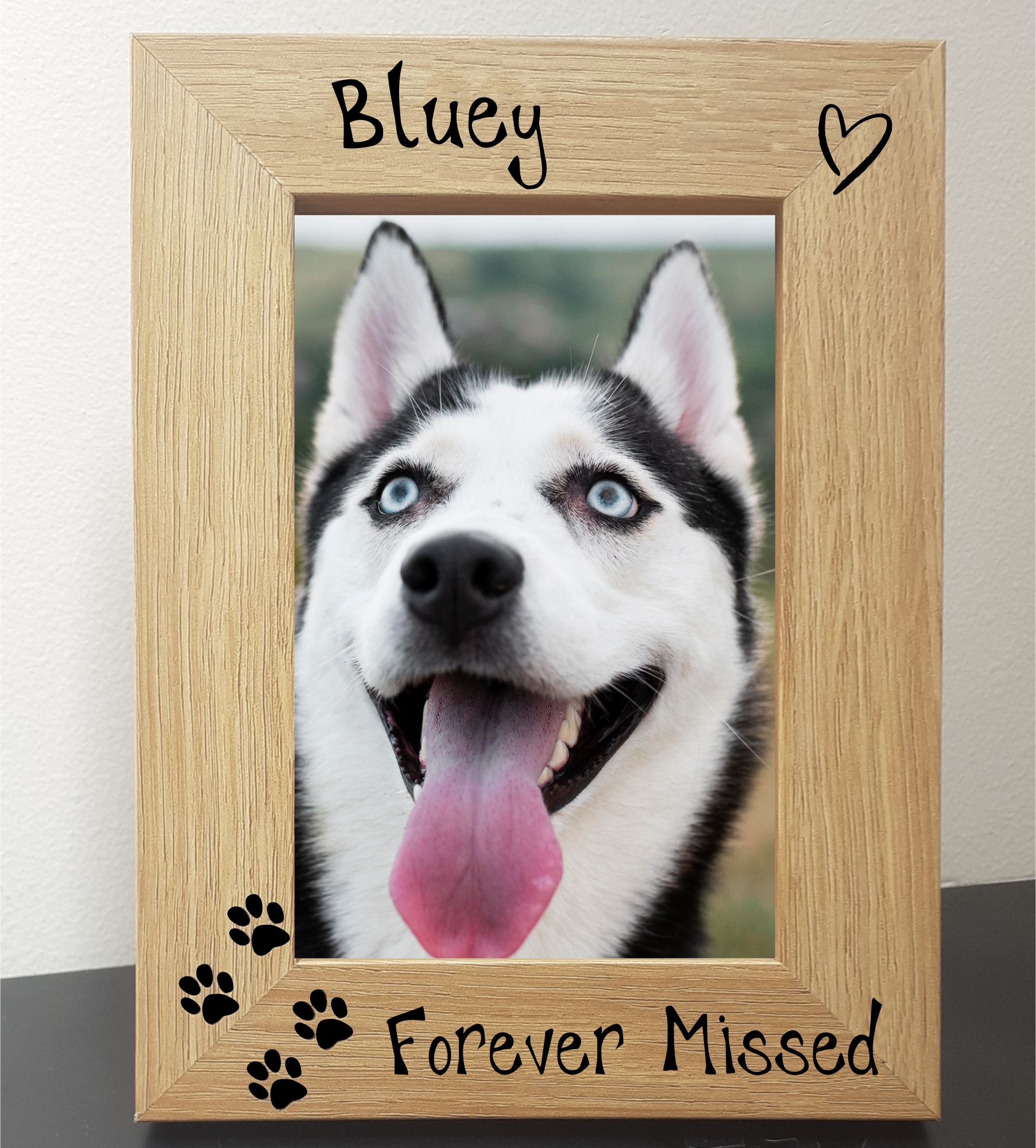 An oak veneer photograph frame. It has been personalised with text etched on the frame, with the dog’s name at the top of the frame, forever missed at the bottom, 4 paw prints etched in the bottom left-hand side of the frame, a heart top right