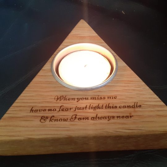 Personalised Solid Oak Wooden Candleholder For Memory Tribute, Triangle-shaped solid oak candleholder with laser-engraved message 'When you miss me have no fear, just light this candle and know I will be near'. The holder features a lit candle, casting a warm glow, against a black background.