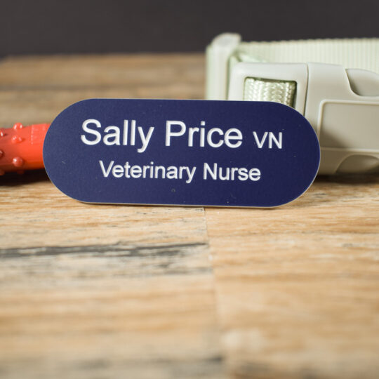 Navy Blue with white text in the shape of a pill name badges, various colours available. The badge is on a wooden background, along with a Collar and a lead