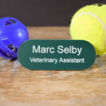 Teal Green with white text in the shape of a pill name badges, line saying Marc Selby and underneath that is Veterinary Assistant. The badge is on a wooden background