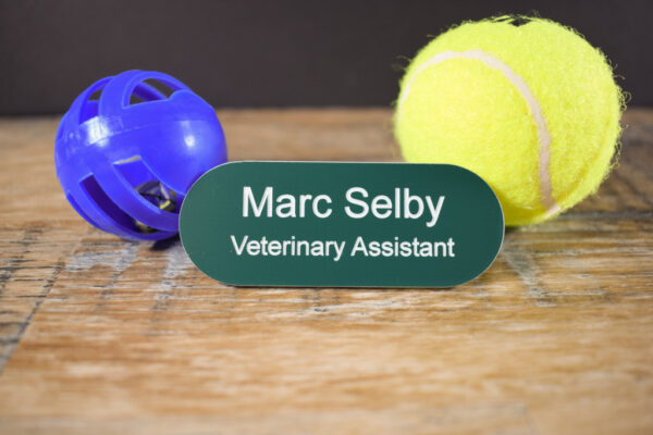 Teal Green with white text in the shape of a pill name badges, line saying Marc Selby and underneath that is Veterinary Assistant. The badge is on a wooden background