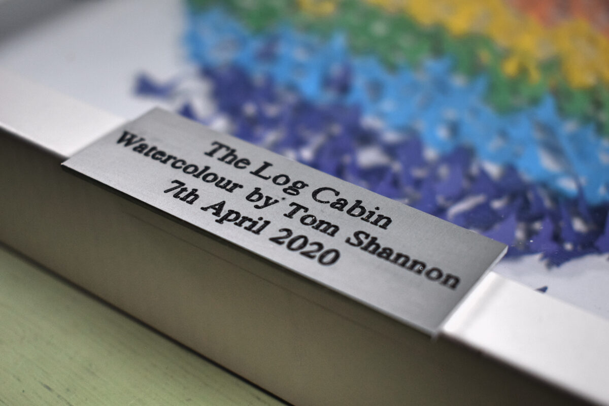 A 75mm x 25mm aluminium plaque with the text 'The Log cabin watercolour by Tom Shannon, 7th April 2020' on a subtle picture frame background