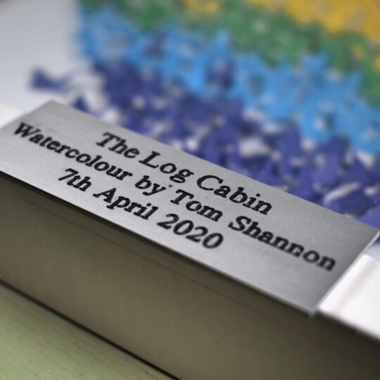 A 75mm x 25mm aluminium plaque with the text 'The Log cabin watercolour by Tom Shannon, 7th April 2020' on a subtle picture frame background