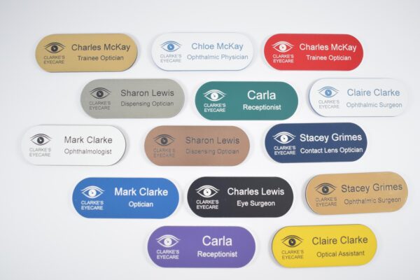 Various colours, pill-shaped name badges have all been engraved. The badges are on a plain white background,