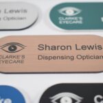 Various colours, pill-shaped name badges with a bronze and black engraving in the centre of the other badges. The badges are on a plain white background,