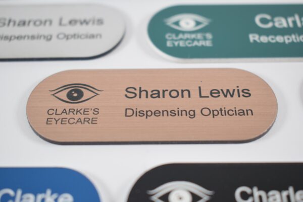 Various colours, pill-shaped name badges with a bronze and black engraving in the centre of the other badges. The badges are on a plain white background,