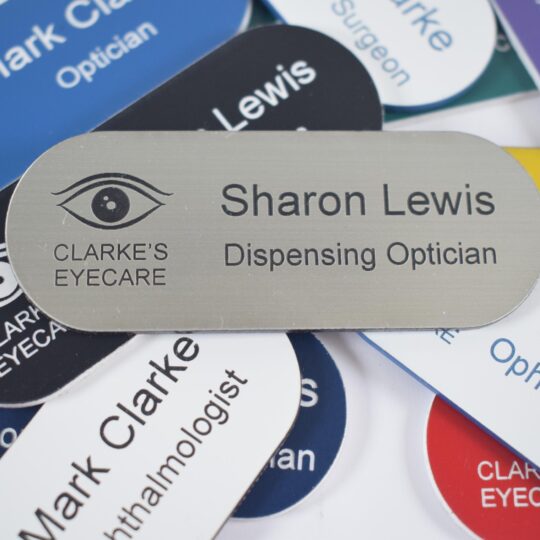 Various colours, pill-shaped name badges with a silver and black engraving and an eye logo to the left of the badge, sit in the centre of the other badges. The badges are on a plain white background,