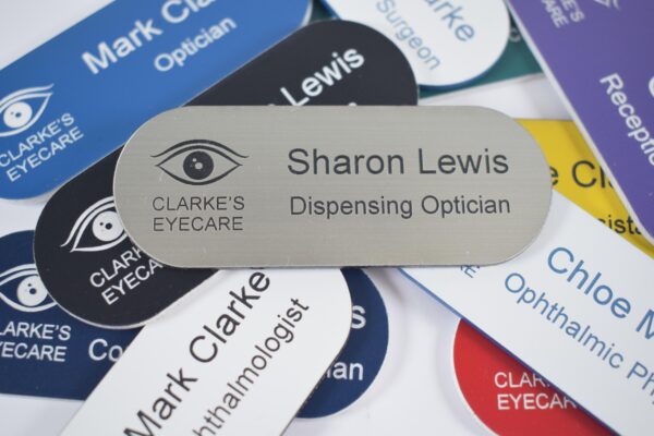 Various colours, pill-shaped name badges with a silver and black engraving and an eye logo to the left of the badge, sit in the centre of the other badges. The badges are on a plain white background,