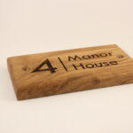 A close-up of a rectangular oak house sign engraved with "4 Manor House" in black lettering, fixed to a textured red-orange brick wall. A vine is growing next to the sign, and a window sill with a potted plant is visible.