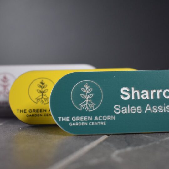 A collection of vibrant and diverse Personalised garden name badges, pill-shaped name badges, each uniquely coloured in shades such as green, gold, silver, blue, and yellow. The badges are designed with a distinctive logo on the left-hand side