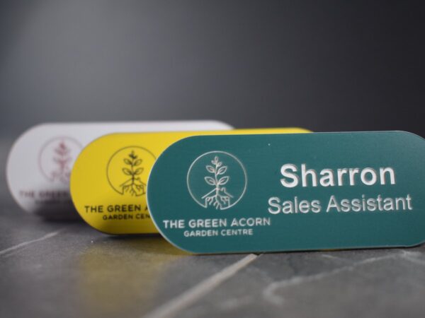 A collection of vibrant and diverse Personalised garden name badges, pill-shaped name badges, each uniquely coloured in shades such as green, gold, silver, blue, and yellow. The badges are designed with a distinctive logo on the left-hand side