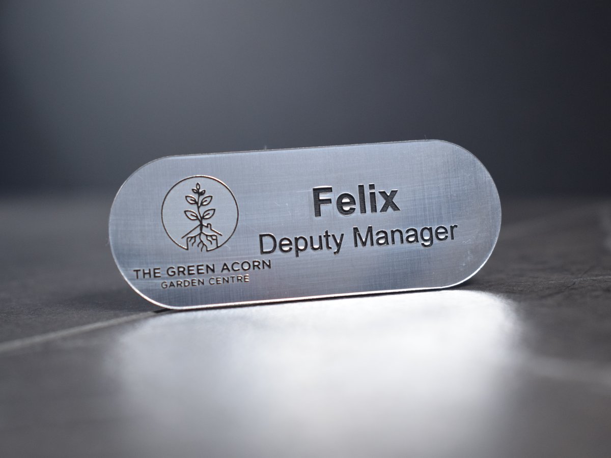 Brushed Silver with black text in the shape of a pill, Logo to the left-hand side of the badge displaying the green acorn garden centre logo. The top line says Felix, underneath a new line saying Manager. The badge is on a clean grey background