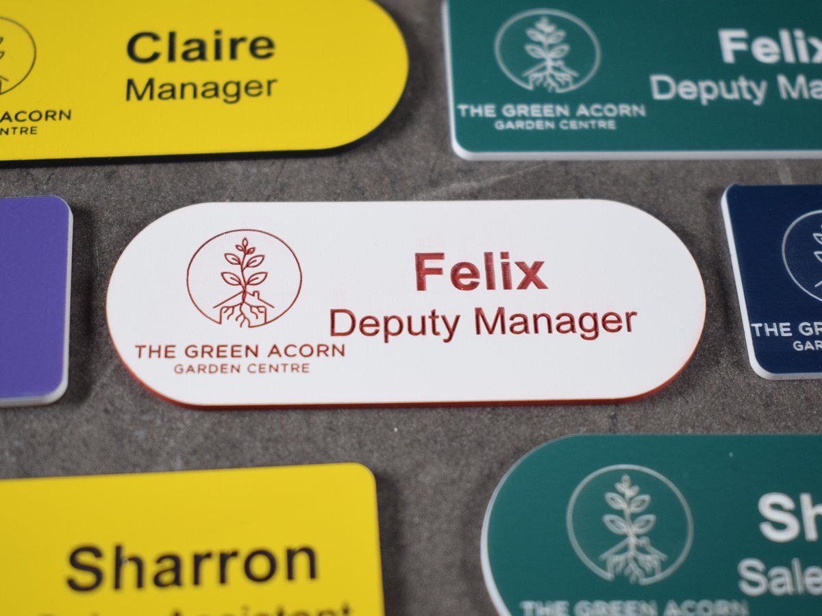Various colours, pill-shaped name badges have all been engraved. The badges are on a plain white background,