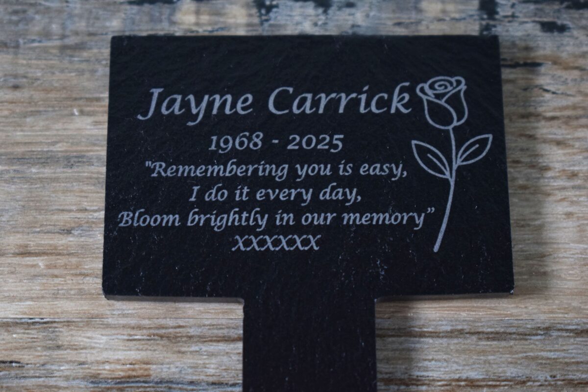 A close-up photograph of a black slate T-shaped pet memorial stake resting on a rustic wooden surface. The plaque is engraved with text and a rose icon on the right-hand side of the plaque. The engraving reads: "Jayne Carrick 1968–2025. "Remembering you is easy, I do it every day, Bloom brightly in our memory” XXXXXX