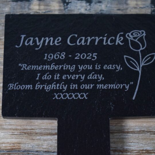 A rectangular black slate memorial garden marker with an integrated spike, featuring a laser-engraved rose on the right. The text reads "Jayne Carrick, 1968 - 2025" followed by the poem "Remembering you is easy, I do it every day, Bloom brightly in our memory." The slate has a natural riven texture and sits on a wooden background.