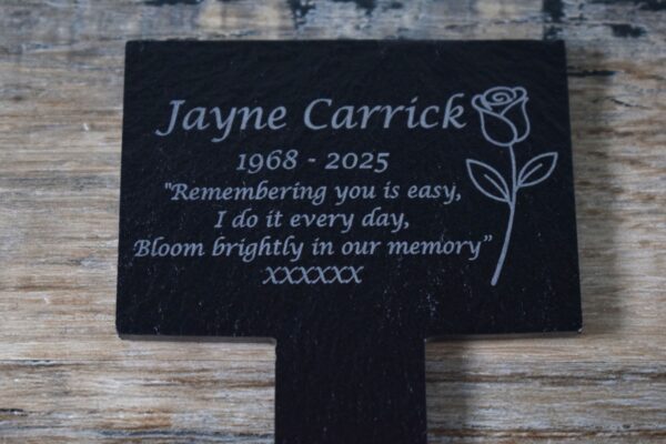 A close-up photograph of a black slate T-shaped pet memorial stake resting on a rustic wooden surface. The plaque is engraved with text and a rose icon on the right-hand side of the plaque. The engraving reads: "Jayne Carrick 1968–2025. "Remembering you is easy, I do it every day, Bloom brightly in our memory” XXXXXX